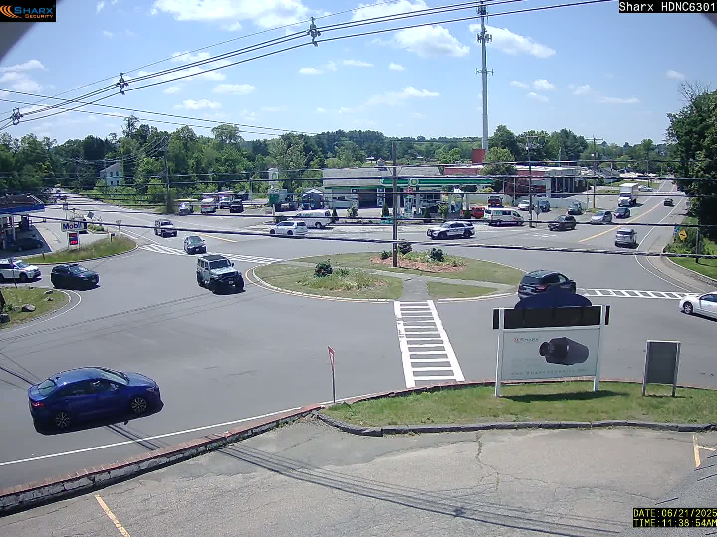 A sunny day shows several cars navigating a roundabout near a gas station and other businesses, surrounded by green trees.