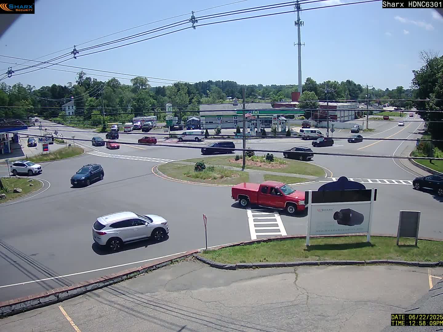 Under a sunny sky, several cars are driving around a traffic circle near a gas station and other buildings.