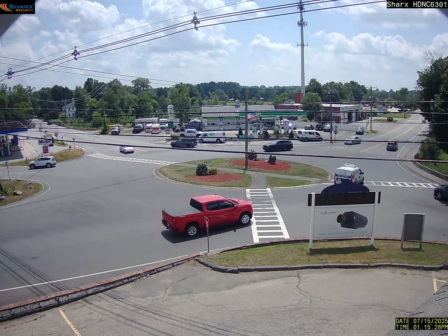 A partly cloudy day shows a red pickup truck and several other vehicles at a roundabout intersection near a gas station and other buildings.