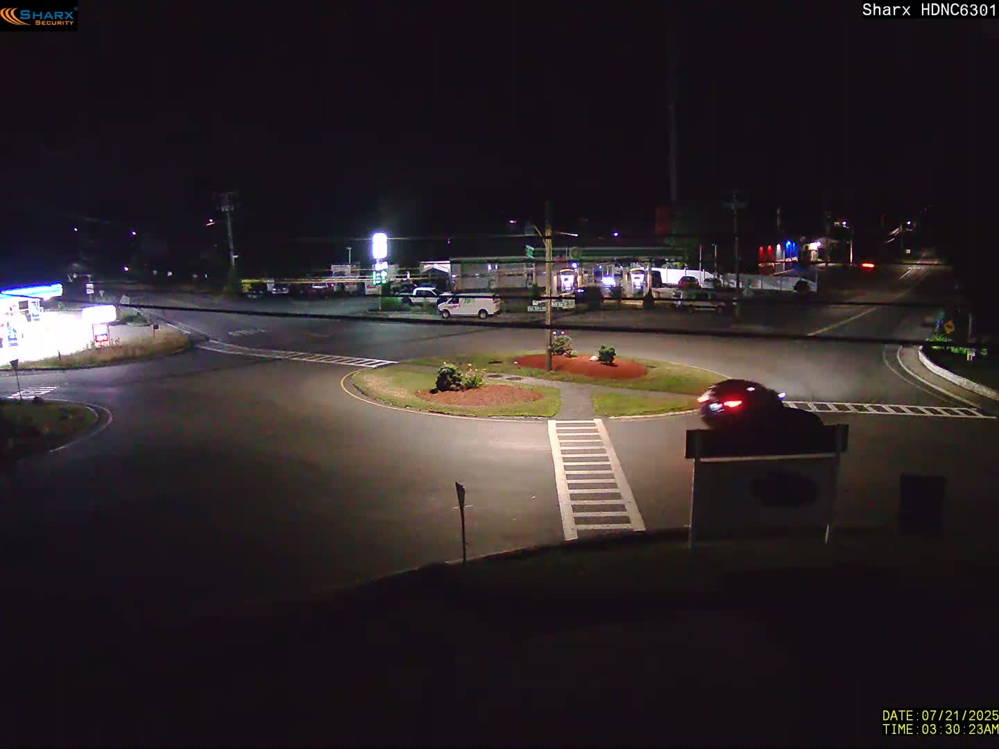 A dark car drives through a roundabout at night, near a gas station and other businesses under artificial light.