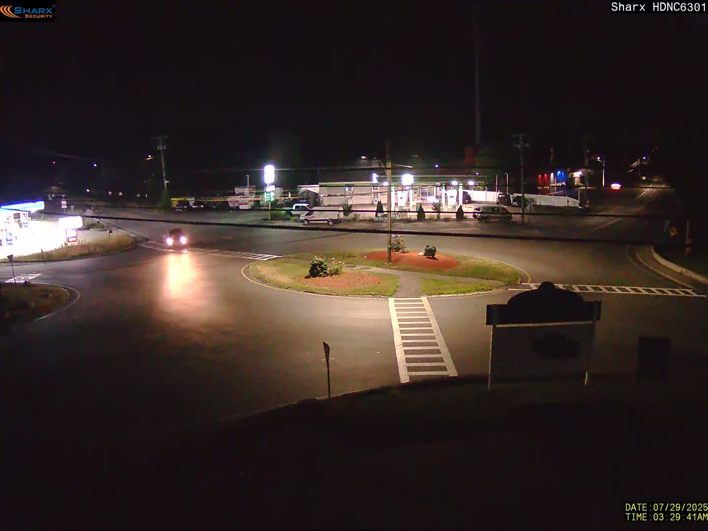 A nighttime view of a roundabout near a gas station and other businesses, with one car visible in the roundabout.