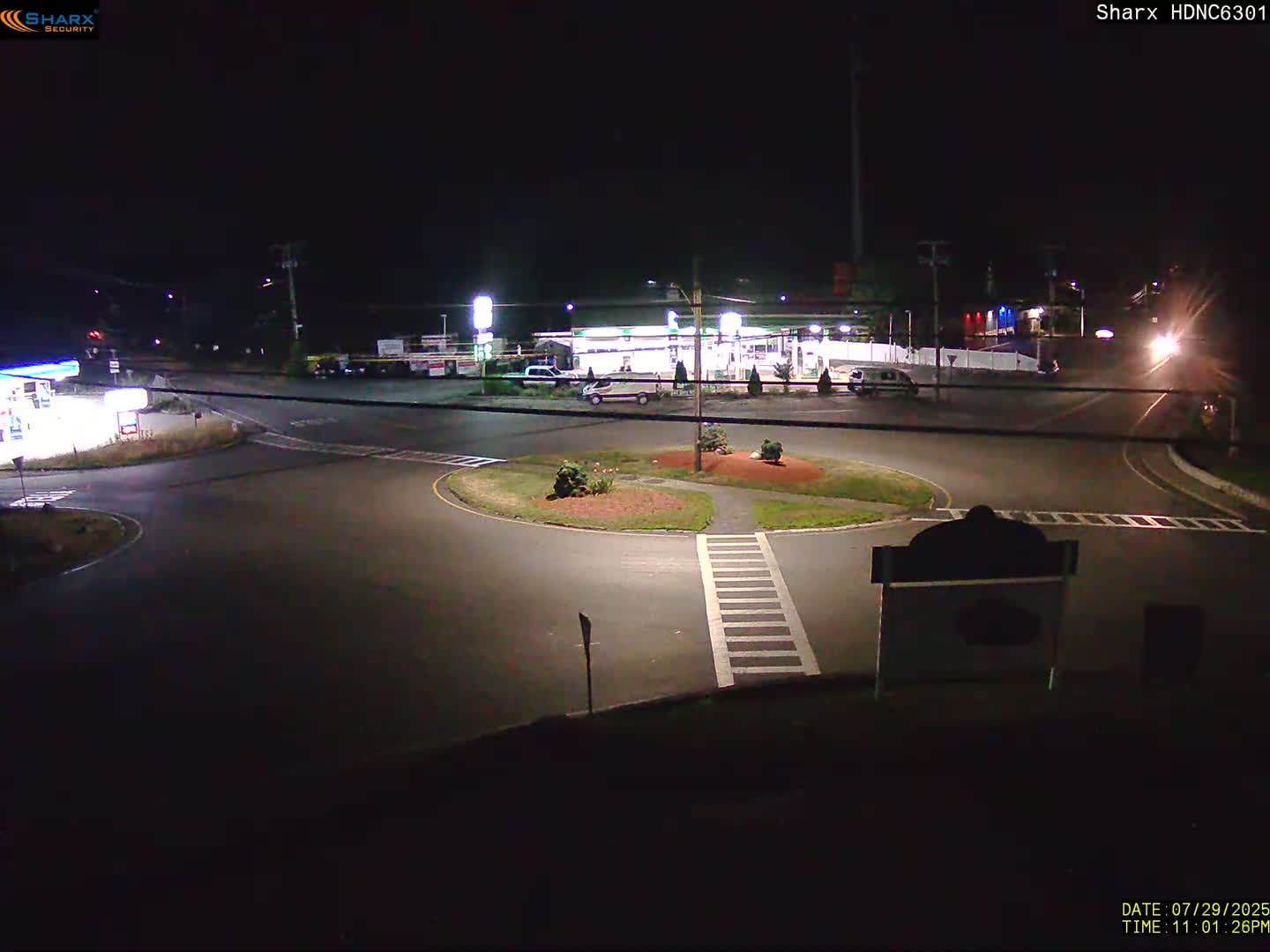 A nighttime view of a roundabout near a gas station and other businesses, with one car visible in the roundabout.