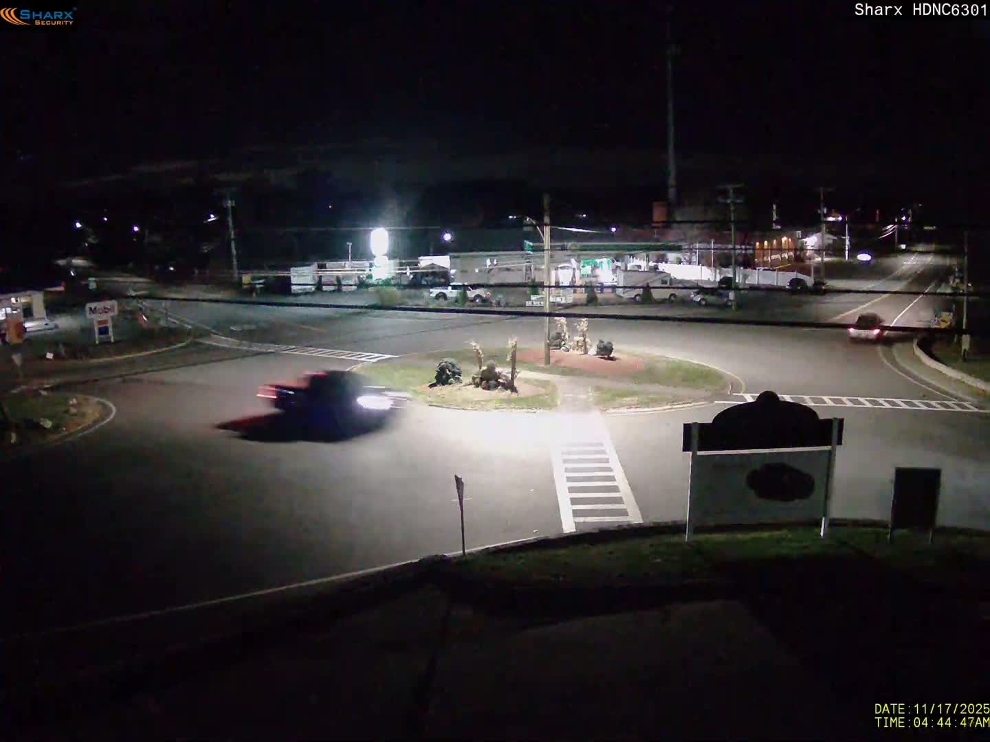 A clear night view shows a motion-blurred car with bright headlights navigating a traffic circle at a road intersection, surrounded by streetlights and distant commercial buildings.