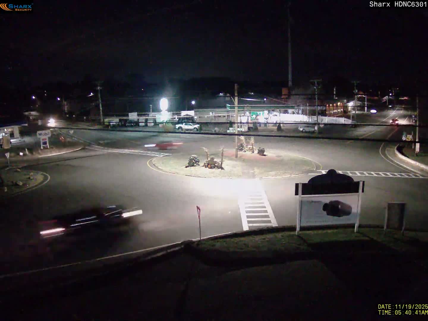 A clear night view shows a motion-blurred car with bright headlights navigating a traffic circle at a road intersection, surrounded by streetlights and distant commercial buildings.