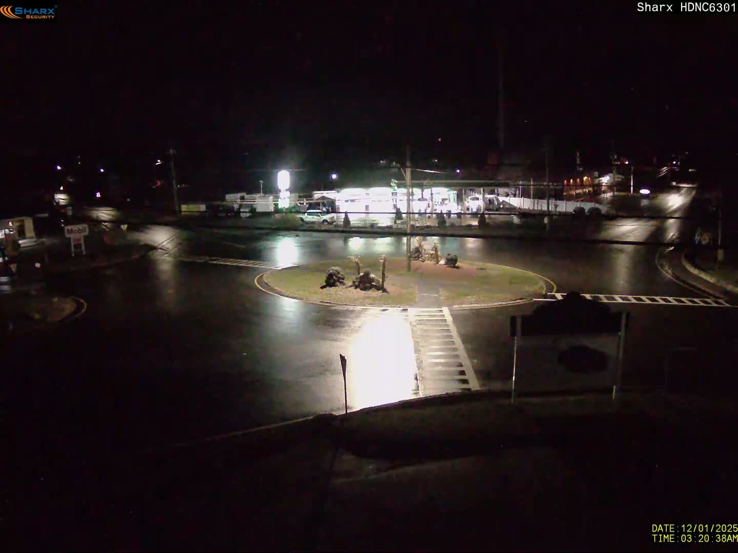 A nighttime view shows a wet roundabout and roads illuminated by streetlights and a brightly lit gas station in the background, indicating recent rain under clear skies.
