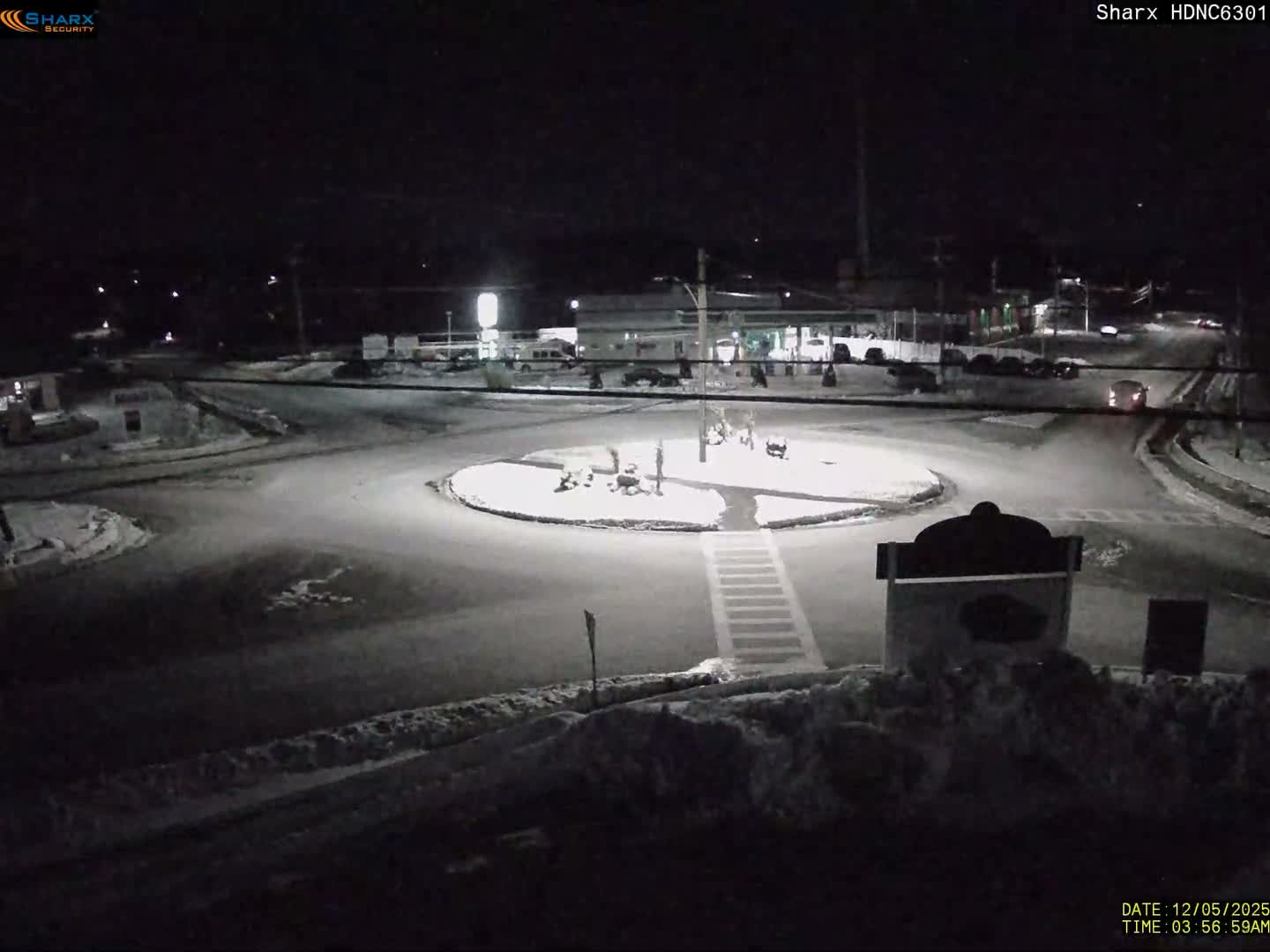 A nighttime scene displays a snow-covered urban roundabout and surrounding streets, illuminated by artificial lights under clear weather conditions.