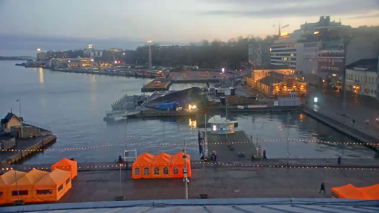 An overcast evening scene shows a harbor with boats, buildings, and orange tents on a waterfront plaza.
