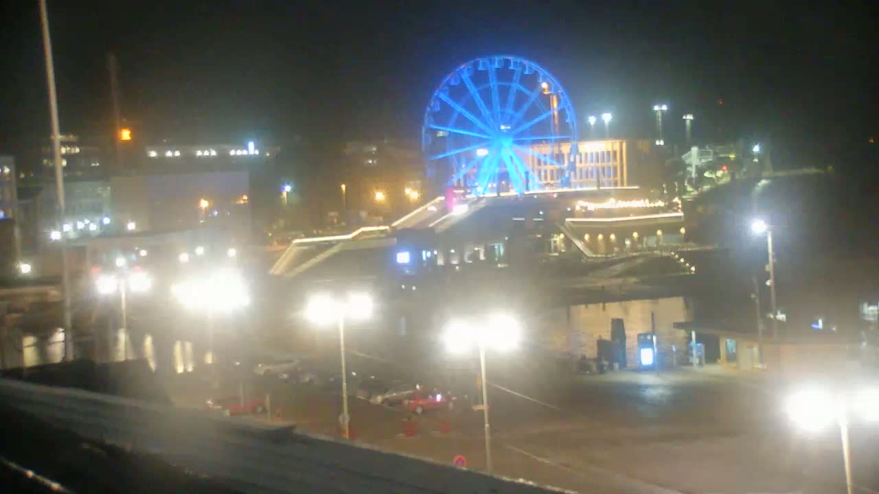 A brightly lit Ferris wheel glows blue against a night sky over a waterfront area with numerous streetlights illuminating parked cars and buildings.