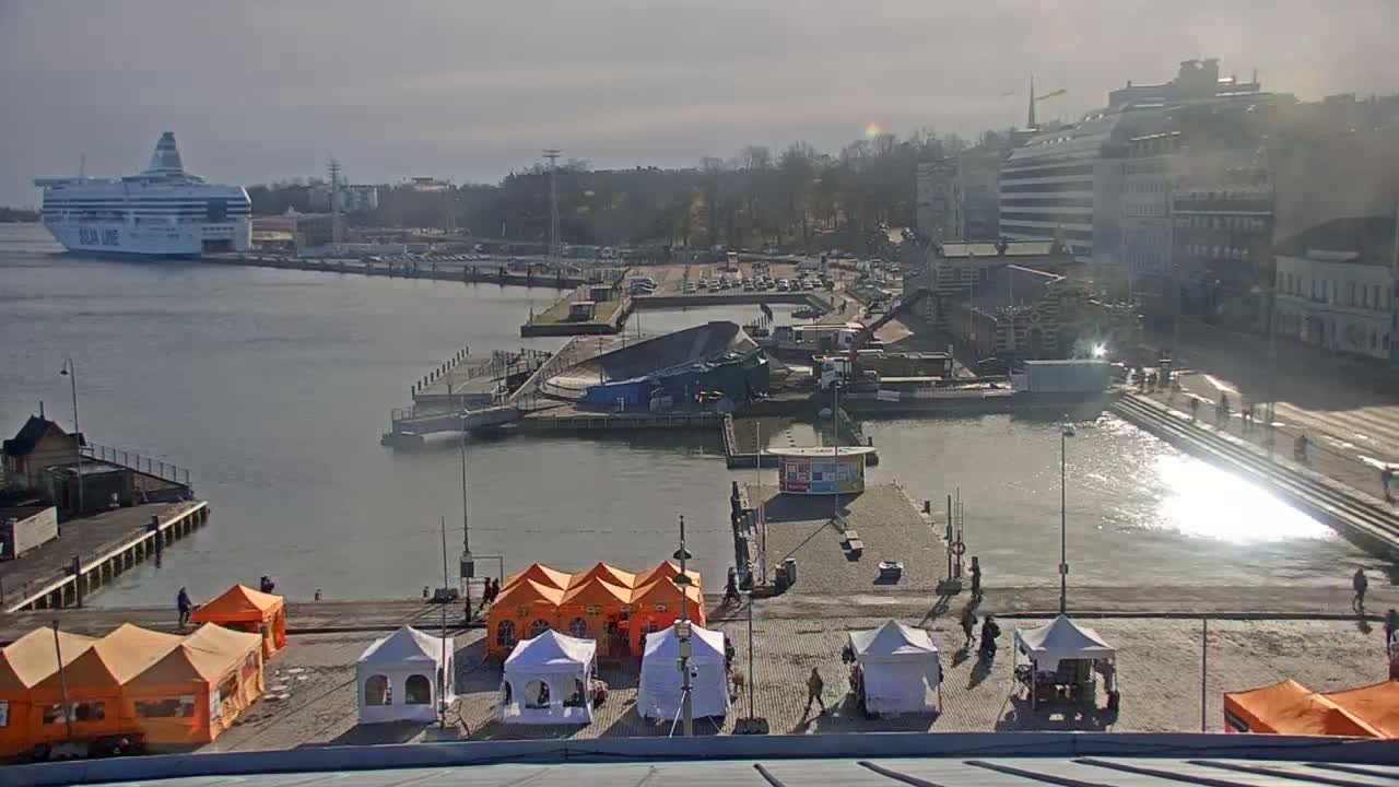 A harbor scene on a sunny day shows a ferry docked, construction activity near a pier, and several tents set up on a waterfront plaza.