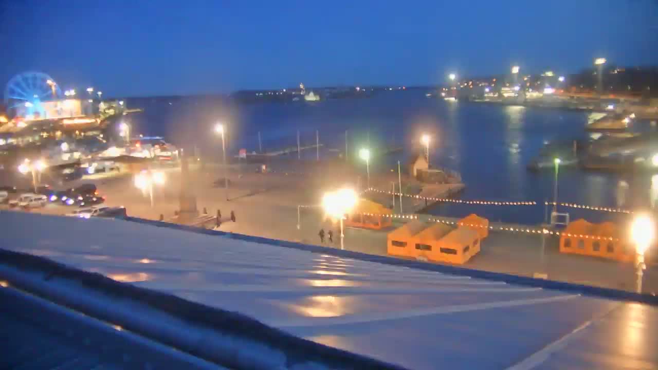 A nighttime view of a harbor with a Ferris wheel, illuminated buildings, and several small orange structures near the water's edge.
