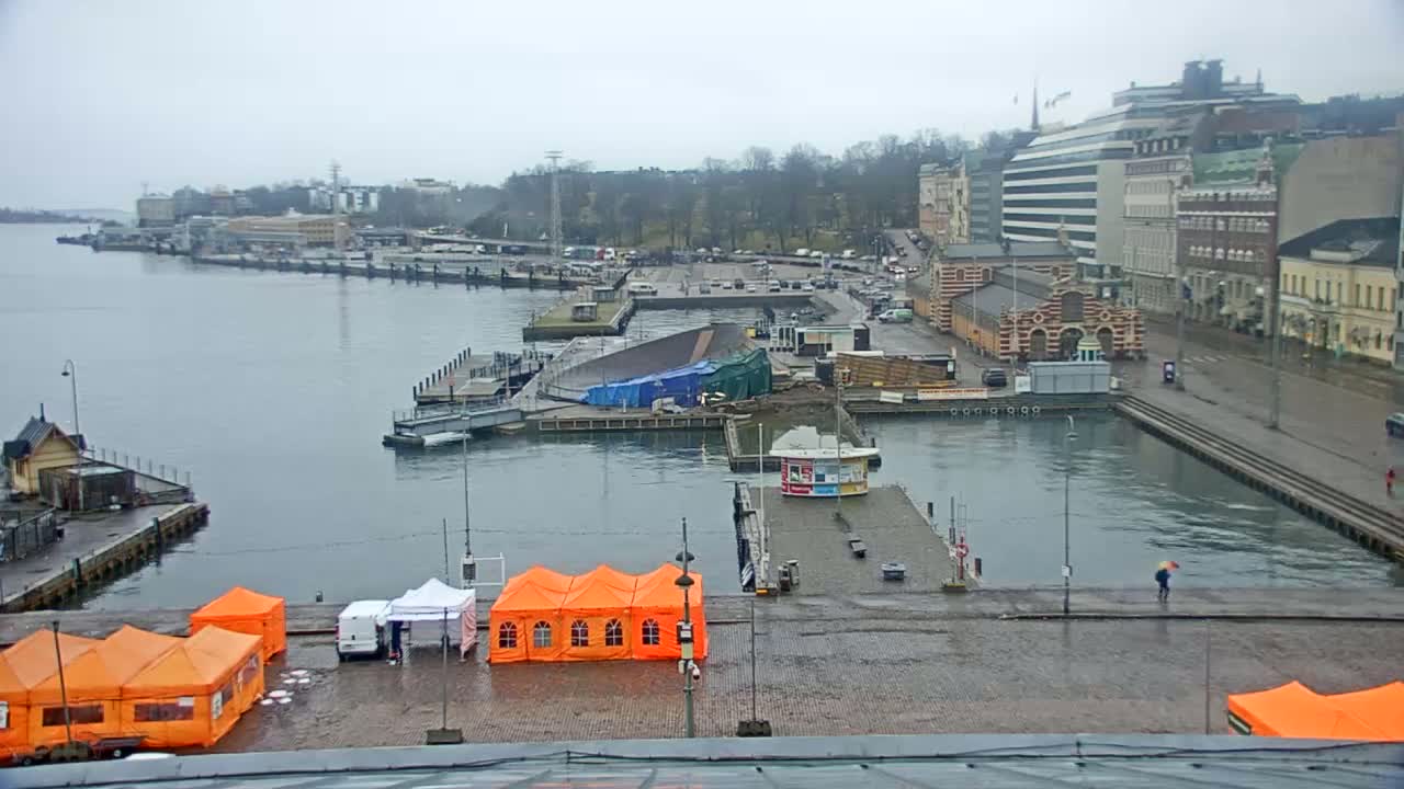 A harbor scene on an overcast day shows a body of water with docked boats, buildings lining the waterfront, and several orange tents set up on the quay.