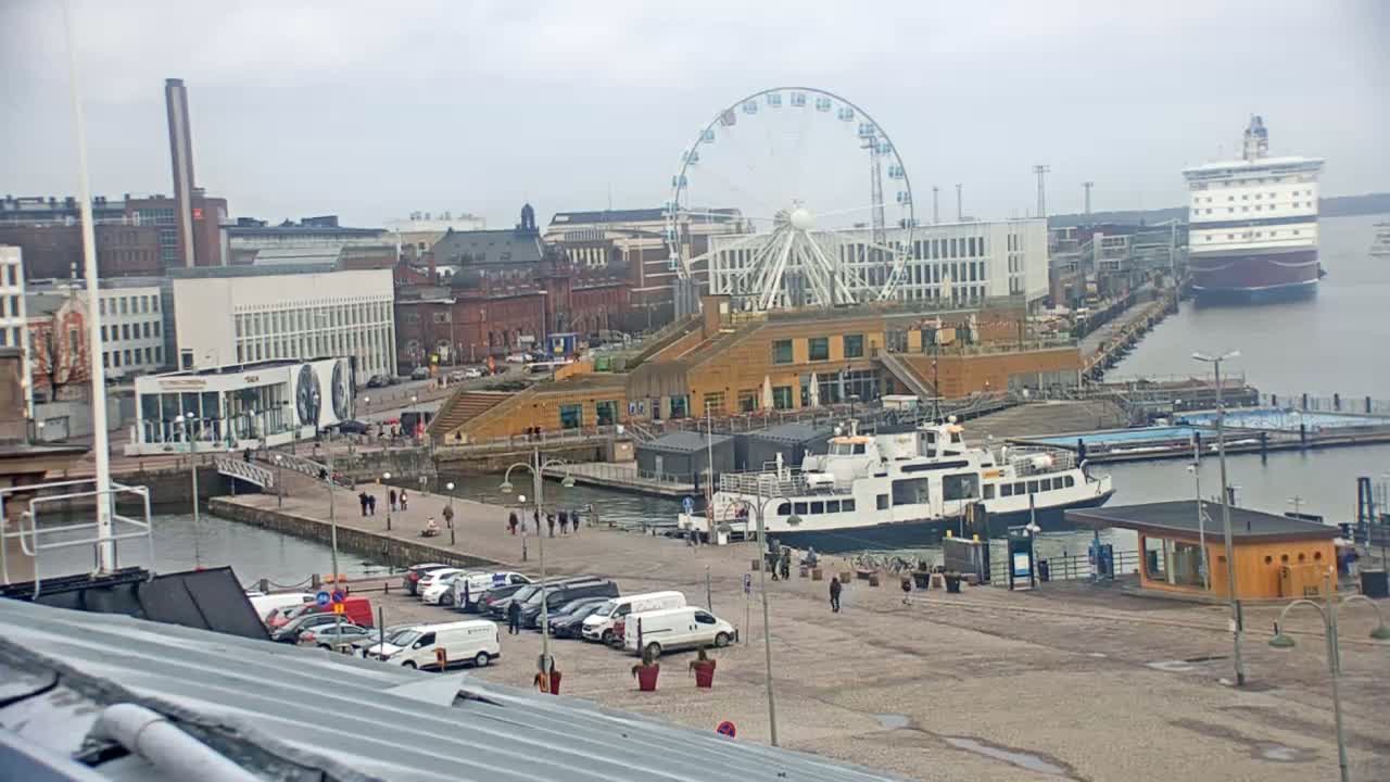 A harbor scene on an overcast day features a Ferris wheel, buildings, docked ferries, and a large ship in the distance.