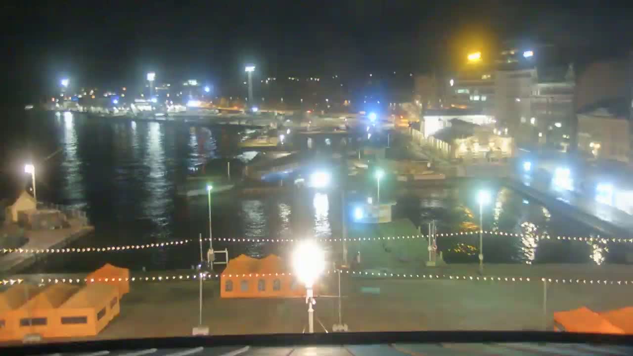 A nighttime view of a harbor with boats moored, illuminated buildings along the shoreline, and orange tents set up on a waterfront plaza.