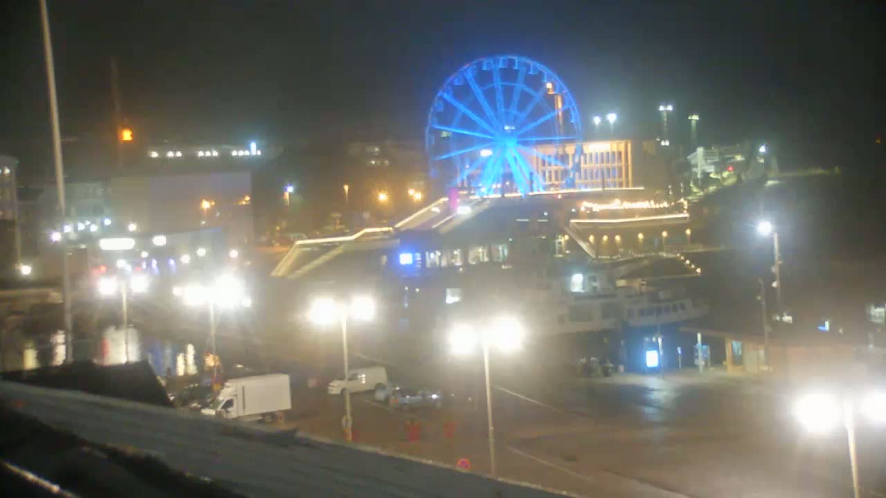 A brightly lit Ferris wheel, illuminated in blue, stands near a waterfront at night, with various vehicles and buildings visible under the lights.