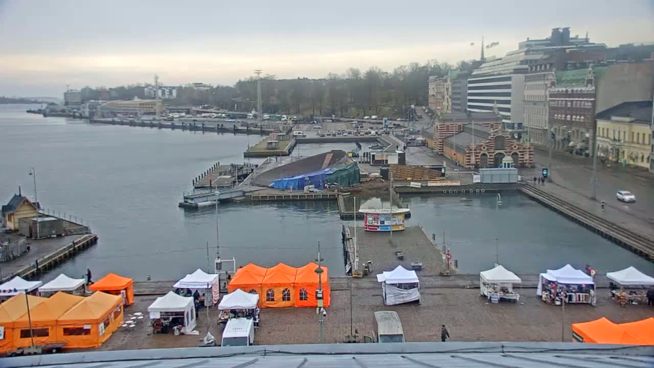 A harbor scene on an overcast day shows market stalls and buildings along a waterfront, with a partially covered structure in the water.