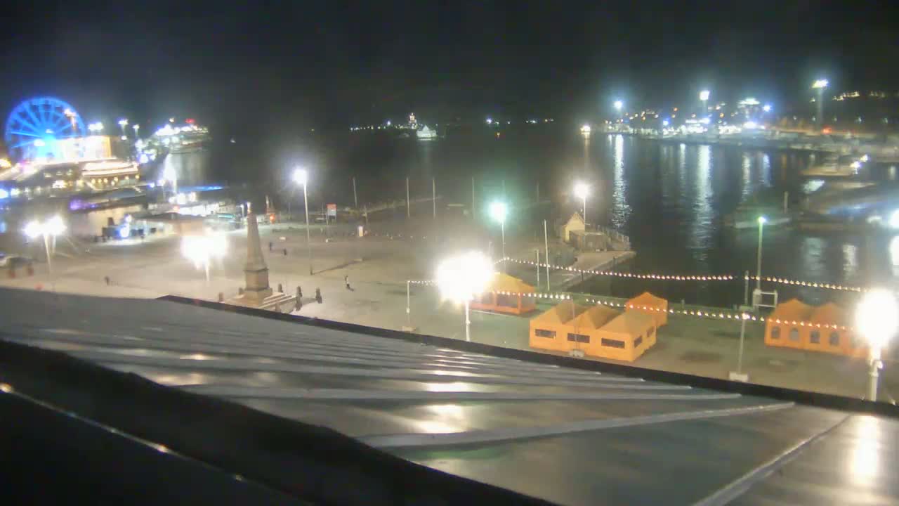 A nighttime view of a harbor with a Ferris wheel, boats, and illuminated structures, overlooking a waterfront plaza from a sloped rooftop.