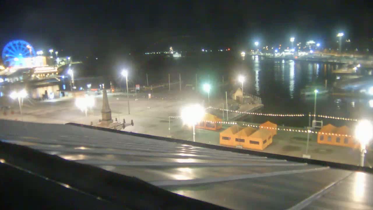 A nighttime view from a rooftop shows a brightly lit waterfront area with a Ferris wheel, a monument, small orange buildings, and a body of water with boats.