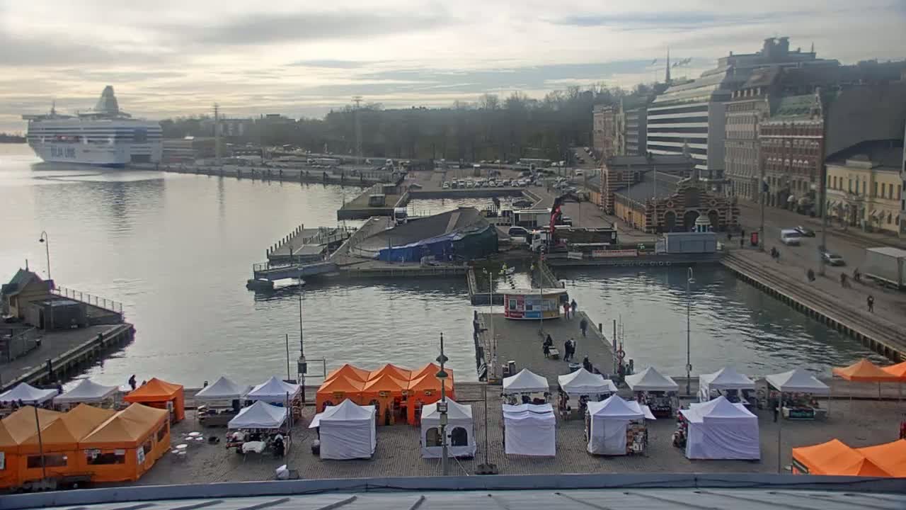 A harbor scene on an overcast day shows a ferry in the distance, a market with numerous white and orange tents in the foreground, and buildings along the waterfront.