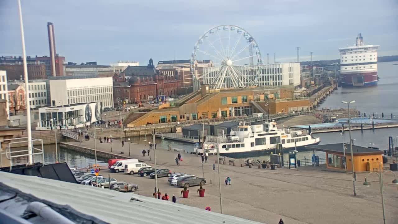 A harbor scene on a cloudy day shows a Ferris wheel, buildings, parked cars, a ferry, and a large ship in the distance.