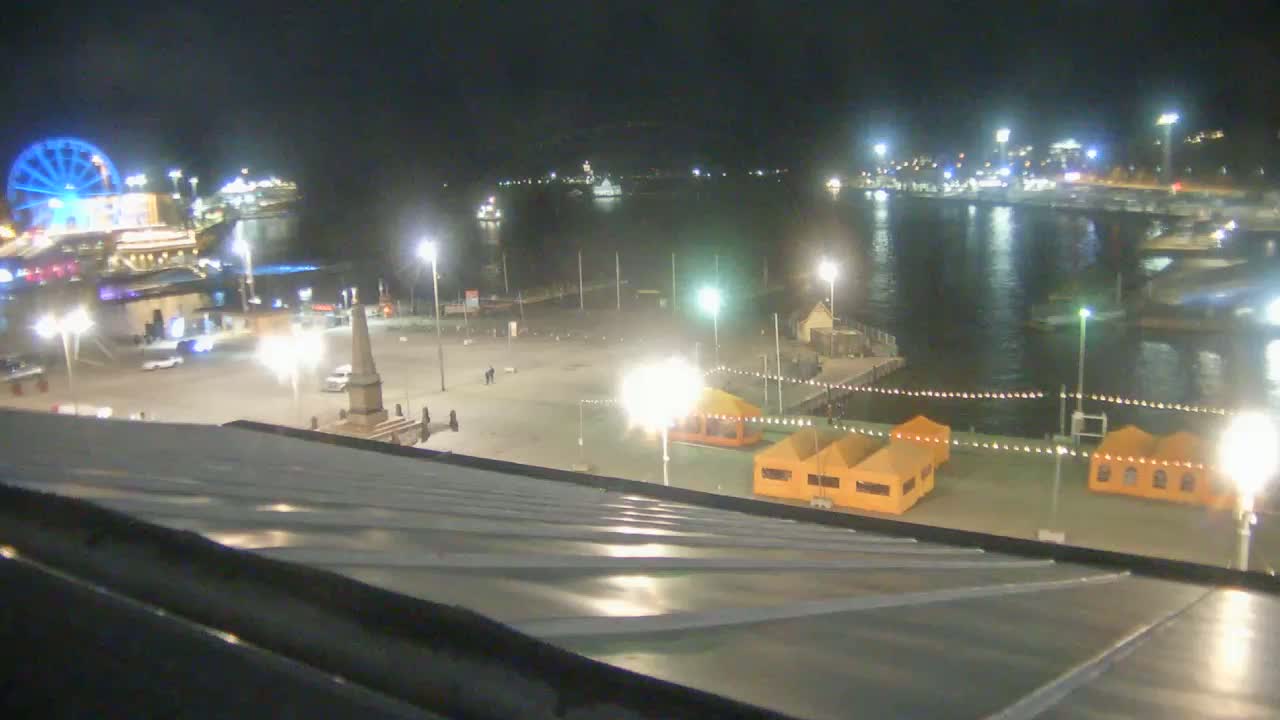 A nighttime view from a rooftop shows a harbor with boats, a Ferris wheel, and a plaza with several illuminated temporary structures.