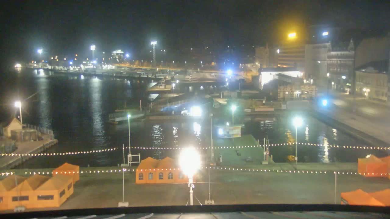 A nighttime view of a harbor with boats, buildings, and orange tents illuminated by numerous lights.