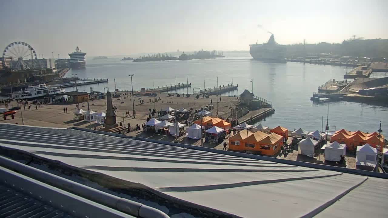 A harbor scene on a hazy, sunny day features a ferry, a large cruise ship in the distance, a Ferris wheel, numerous market stalls, and a pier with people walking around.