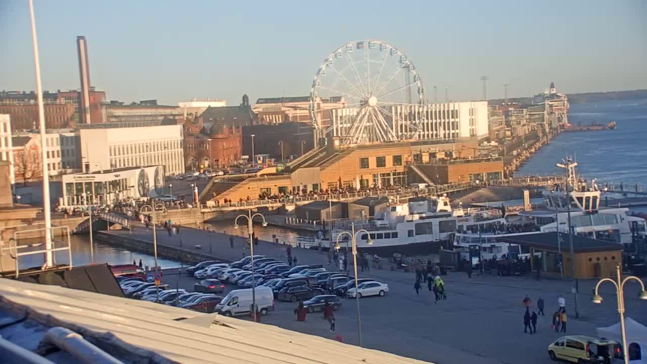 A Ferris wheel overlooks a harbor filled with boats and cars parked along a waterfront promenade under a clear, sunny sky.