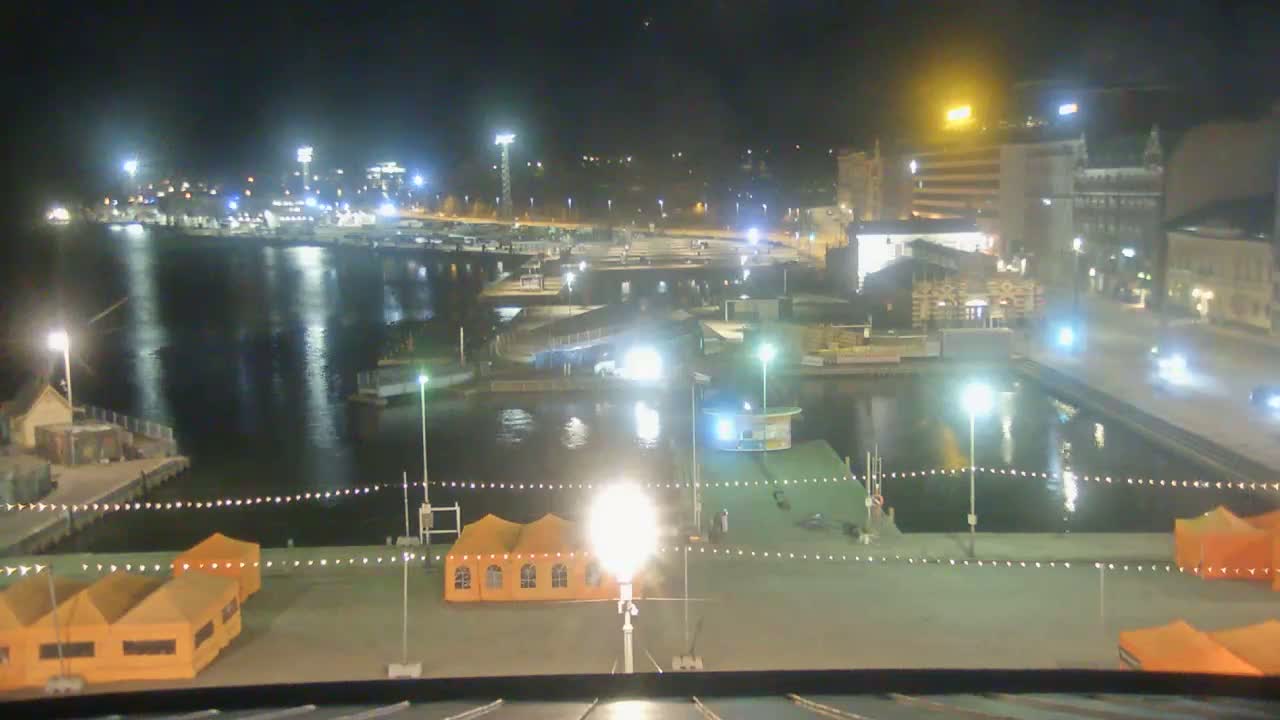 A nighttime view of a harbor with boats docked, buildings in the background, and several orange tents set up on a paved area near the water.