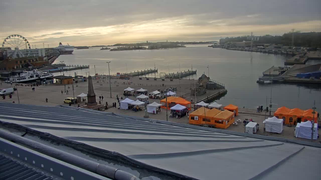 An overcast harbor scene shows a waterfront market with several white and orange tents, a Ferris wheel, docked ferries, and a large body of water.