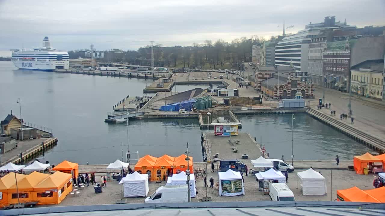 An overcast day at a harbor shows a docked ferry, a market with orange and white tents, and buildings along a waterfront.