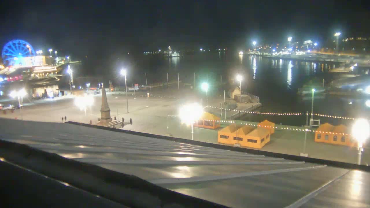 A nighttime view of a harbor and city square, illuminated by numerous lights, featuring a Ferris wheel, a monument, and several small structures along the waterfront under a clear sky.