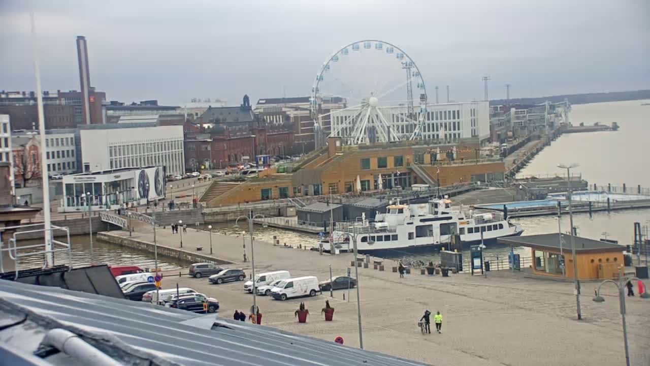 A Ferris wheel overlooks a harbor scene with a ferry docked, several buildings, and a mostly cloudy sky.