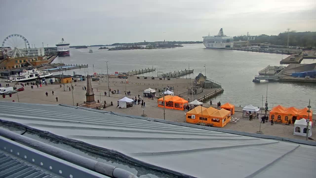 A harbor scene on an overcast day shows a large square with several orange and white market tents, a Ferris wheel, and numerous ships in the water.