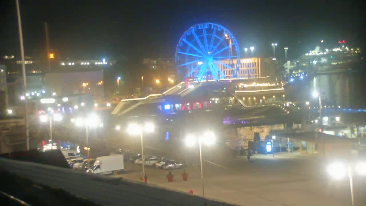 A brightly lit Ferris wheel is prominent in a nighttime cityscape, with cars parked in a lot below and other buildings and lights visible in the distance.