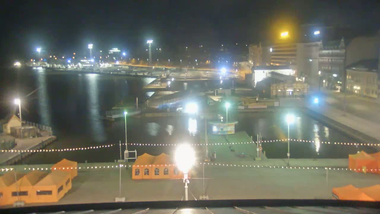 A nighttime view of a harbor with brightly lit buildings, docked boats, and several orange tents along a waterfront area.