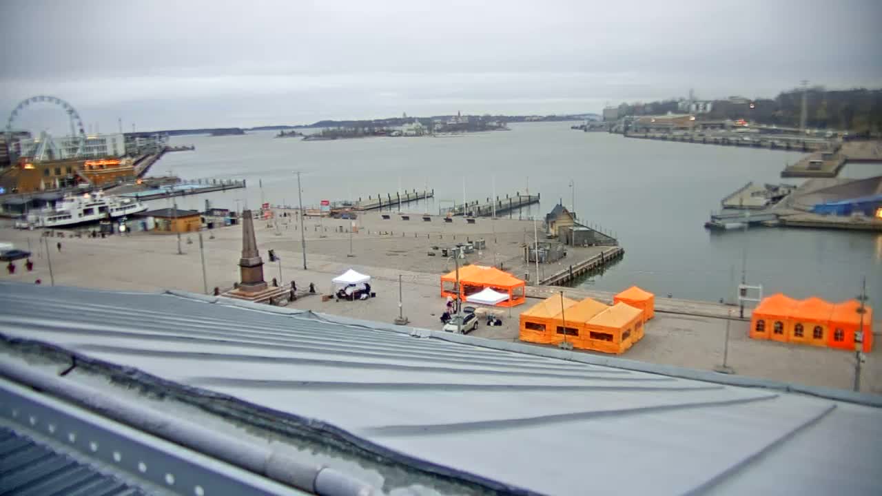 An overcast day reveals a harbor scene with a Ferris wheel, boats, docked piers, several orange and white tents, and a monument in a plaza.