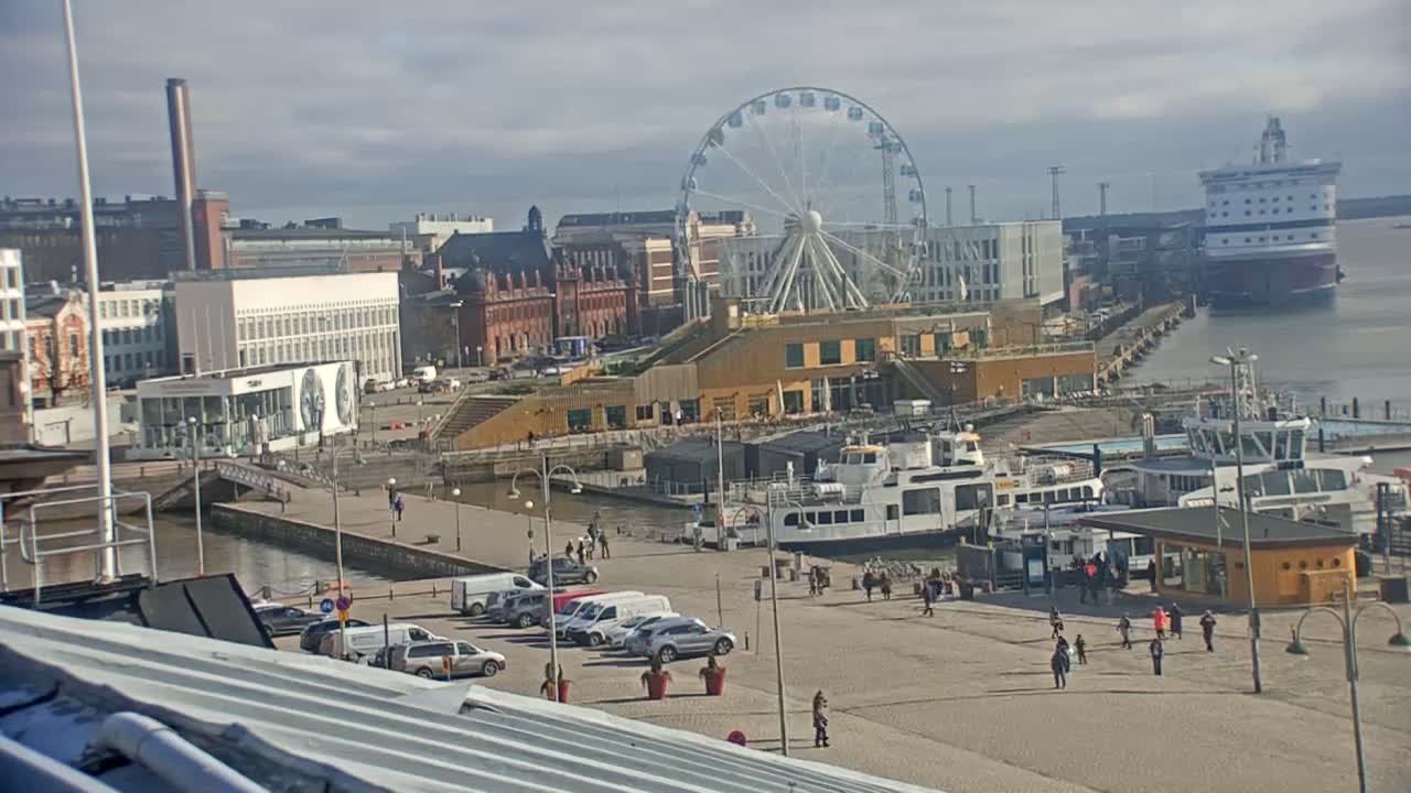 A harbor scene on a partly cloudy day features a Ferris wheel, several docked ferries, a large cruise ship, and a cityscape with various buildings.