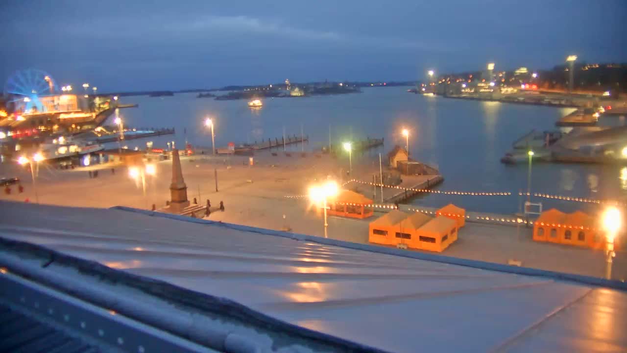 A nighttime view of a harbor illuminated by streetlights, with a Ferris wheel visible in the distance, and orange tents set up near the water's edge.