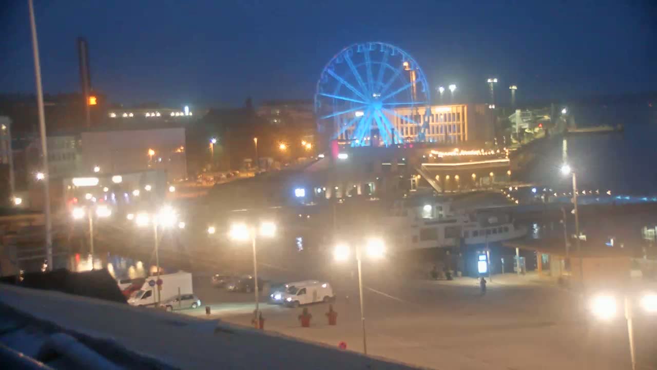 A brightly lit waterfront at night features a Ferris wheel illuminated in blue, buildings, vehicles, and a body of water.