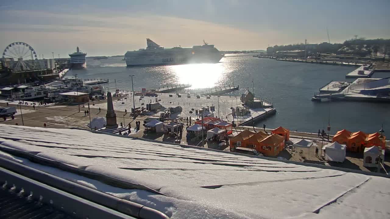 A snow-covered harbor scene on a sunny day shows a large ferry and several smaller boats docked near a Ferris wheel and market stalls.