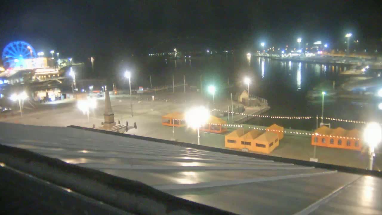 A nighttime view of a waterfront area with a Ferris wheel, illuminated tents, and a calm body of water.