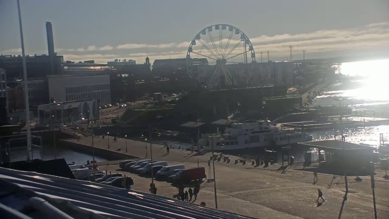 A waterfront cityscape on a sunny day shows a Ferris wheel, buildings, parked cars, a ferry, and people walking along a harbor.