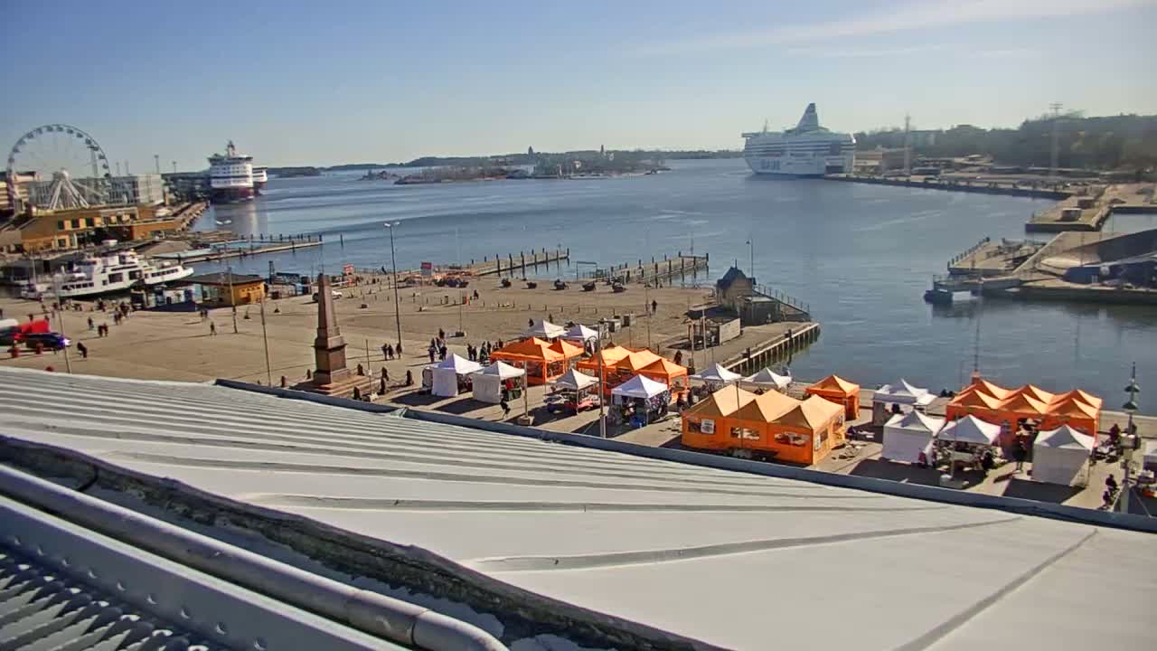A harbor scene on a sunny day shows a Ferris wheel, several ships, and a collection of orange and white market stalls situated near a body of water.