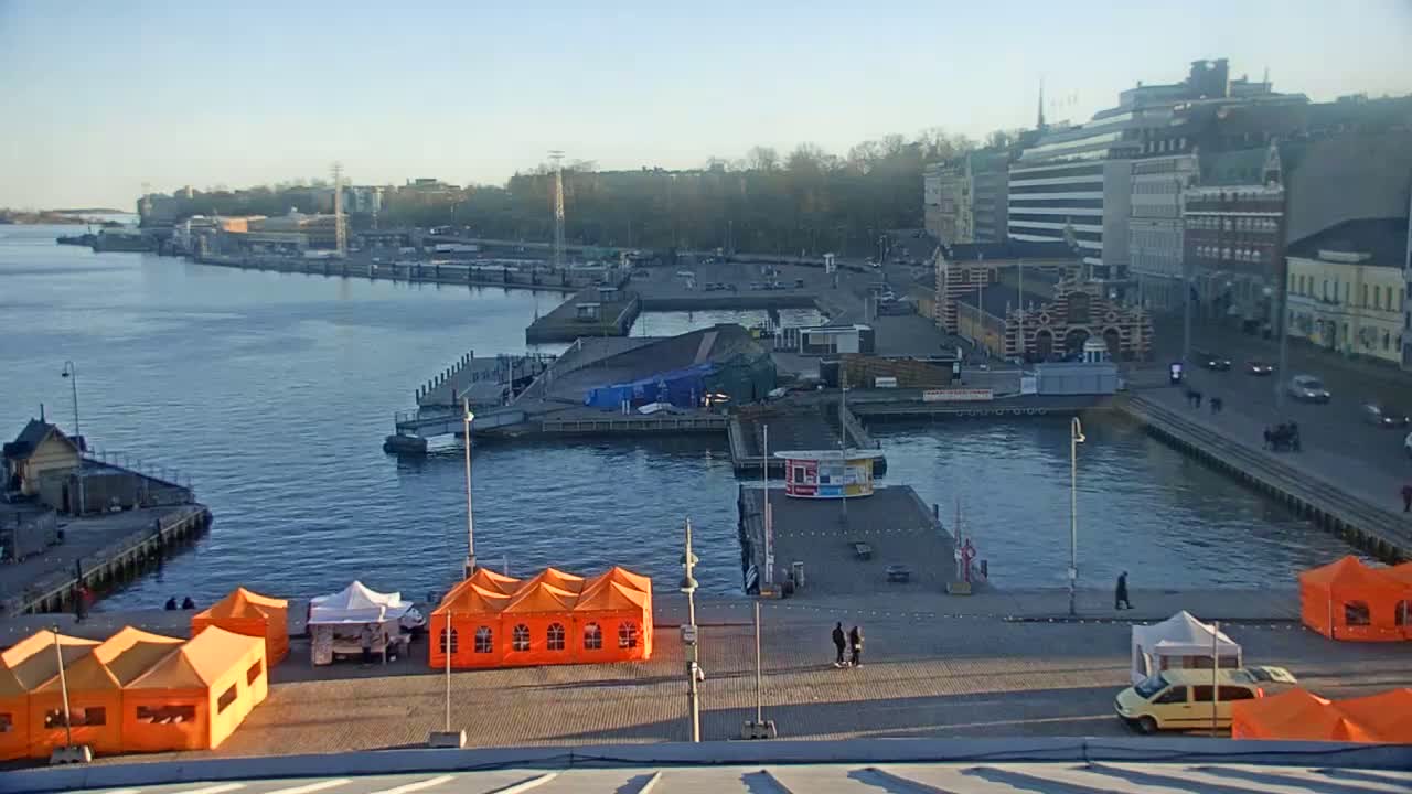 An outdoor harbor scene on a clear, sunny day shows several boats, docks, buildings, and orange tents on a paved area near the water.