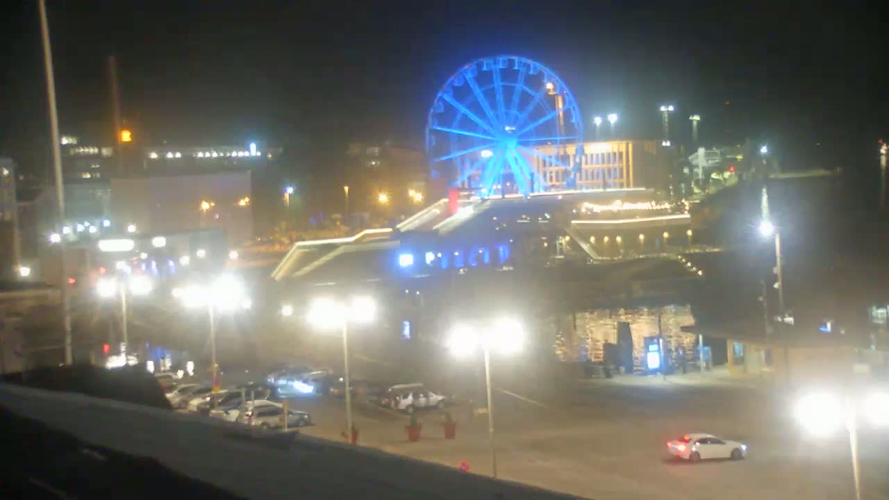 A brightly lit nighttime scene features a large Ferris wheel illuminated in blue, surrounded by buildings, streetlights, and parked cars.