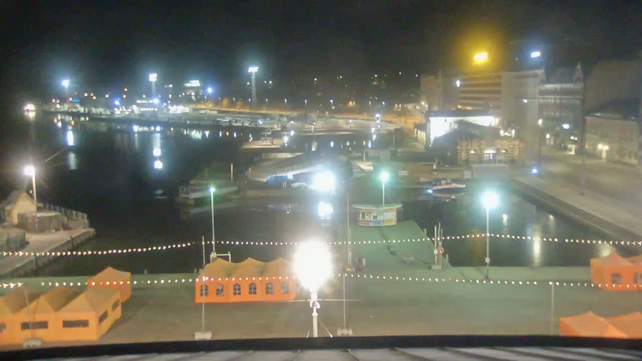 A nighttime view of a harbor with several boats docked, illuminated buildings in the background, and orange tents near the foreground.