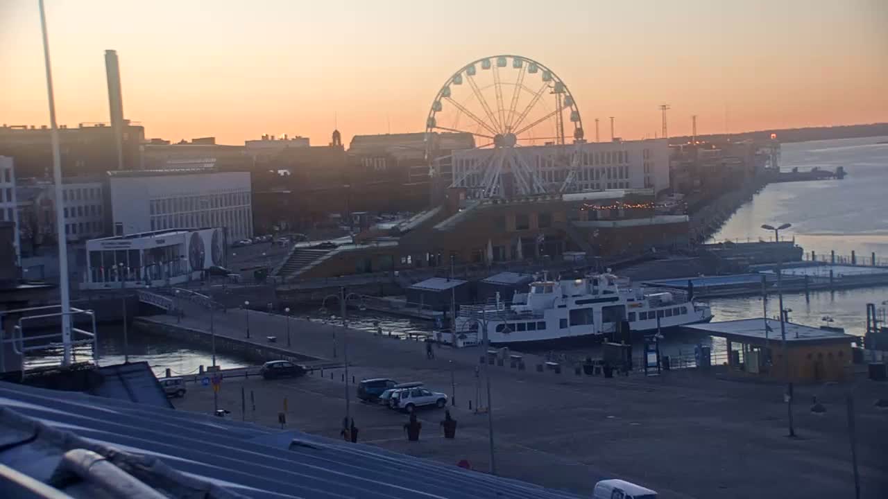 A Ferris wheel overlooks a harbor at sunset, with a ferry docked nearby and several cars parked in a lot.