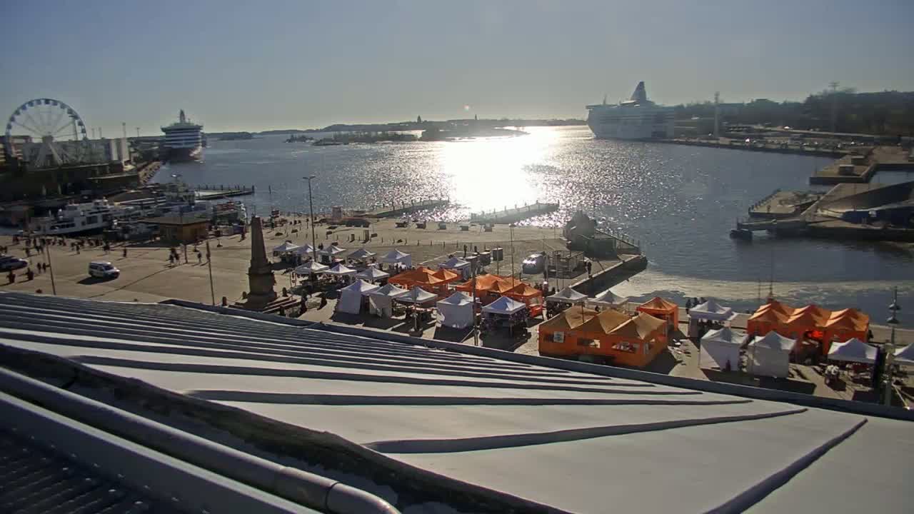 A waterfront market with numerous white and orange tents is situated near a harbor with a ferry and a Ferris wheel under sunny conditions.
