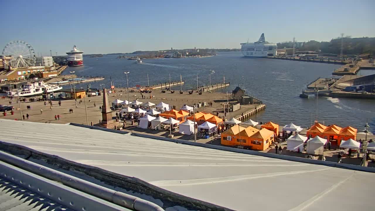 A harbor scene on a sunny day shows a Ferris wheel, several ships, and numerous market stalls set up on a waterfront plaza.