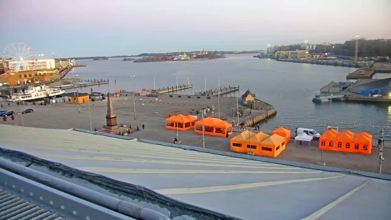 An elevated view of a harbor with several orange tents set up on a plaza near a ferry terminal and a Ferris wheel, under a twilight sky.